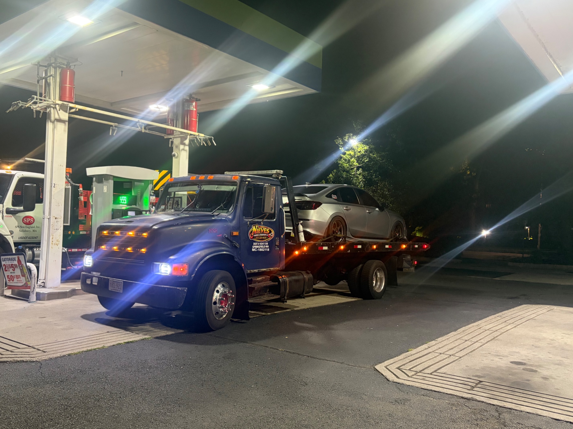 Nieves Towing flatbed with a silver sedan at a gas station at night