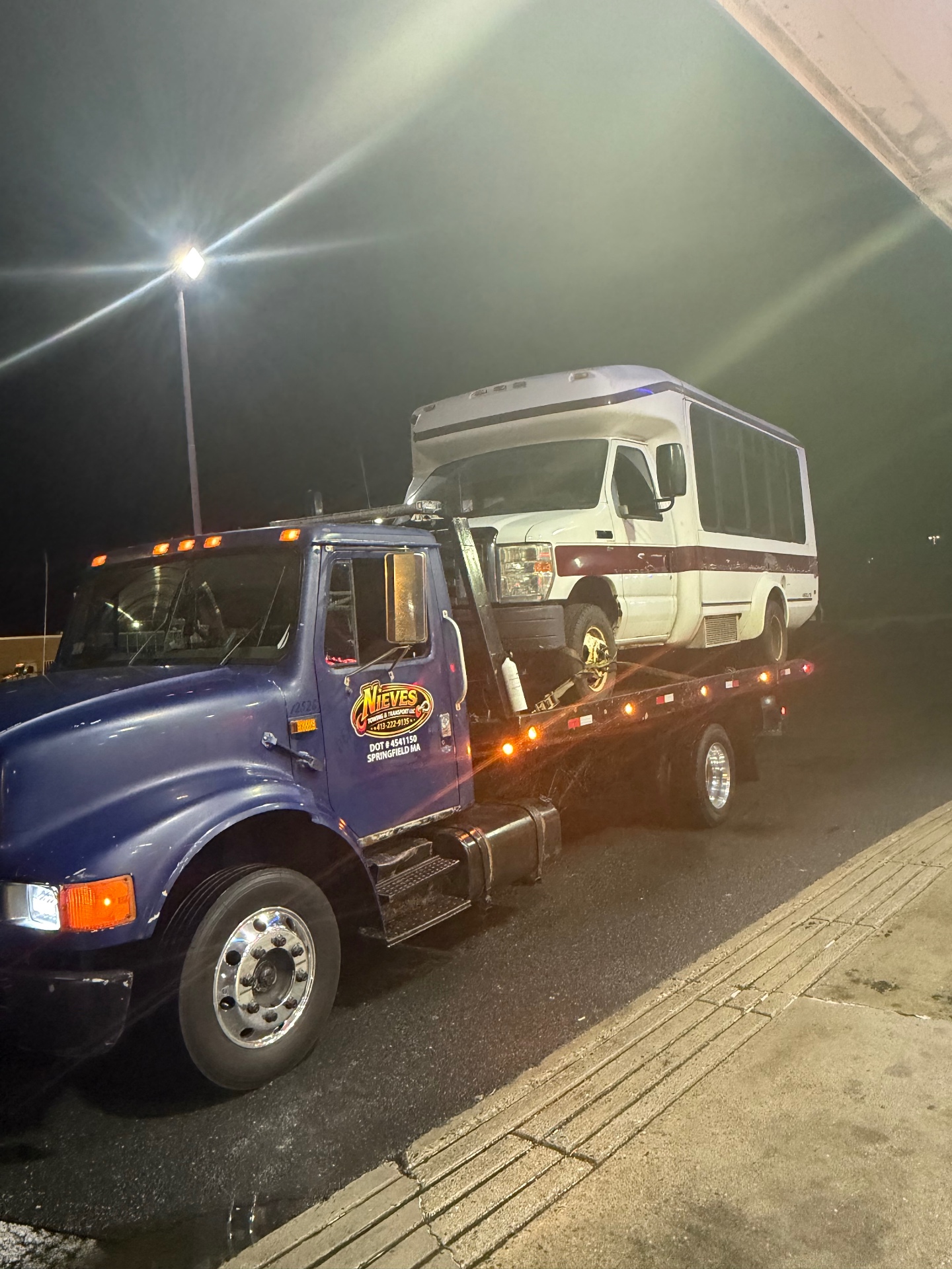 Nieves Towing flatbed hauling a white shuttle bus at night in Springfield, MA